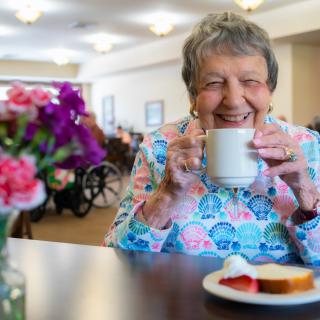 An elderly woman enjoying a cup of coffee with breakfast in a cheerful setting.
