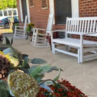 A cozy porch with white benches and rocking chairs beside a brick building, adorned with decorative plants.