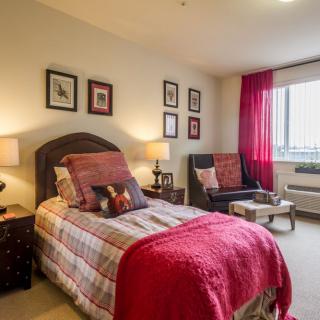 A cozy bedroom with a single bed, adorned with decorative pillows and warm lighting, featuring framed artwork on the walls and a comfortable armchair by the window.