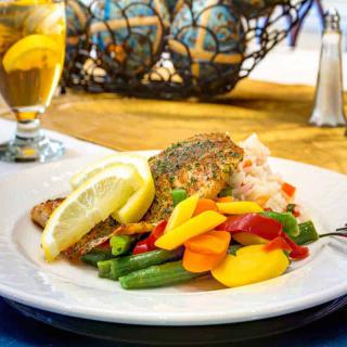 A plate of seasoned fish with colorful vegetables and lemon slices, accompanied by a glass of iced tea.