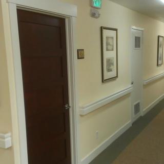 A quiet, well-lit hallway featuring framed pictures and a wooden door with an emergency exit sign above.