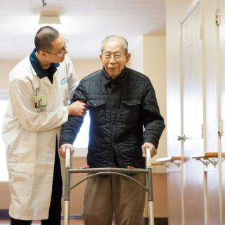 A doctor assists an elderly man using a walker in a hallway.