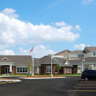 A modern assisted living facility with a well-maintained exterior and parking area under a blue sky.