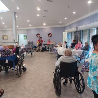 A group of elderly people in a nursing home participating in a social gathering, with some individuals in wheelchairs.