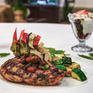A beautifully plated grilled chicken breast served with saut�ed vegetables and a dessert in the background.