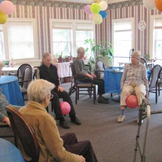 A group of seniors participating in a seated exercise activity with balloons in a community room.