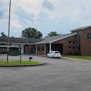 A view of a brick building with a covered entrance and parking area under a cloudy sky.