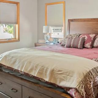 A cozy bedroom with a neatly made bed, featuring decorative pillows and two windows allowing natural light.