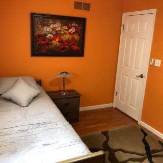 A cozy bedroom with orange walls, featuring a bed, nightstand with a lamp, and a decorative painting.