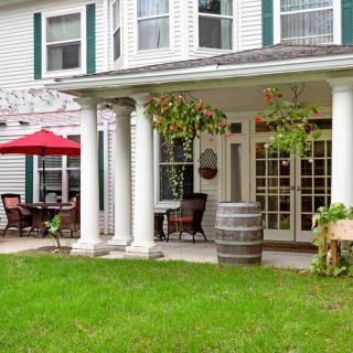 A charming patio with white columns and outdoor seating, featuring a red umbrella and a wooden barrel, set against a lush green lawn.