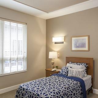 A cozy bedroom with a single bed, decorated in blue and white tones, featuring a window, lamp, and wall art.
