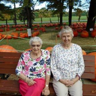 Two elderly women sitting on a bench in a pumpkin patch.