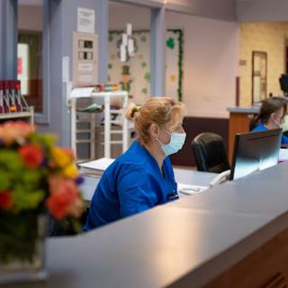 A healthcare receptionist wearing a mask, assisting at the front desk.