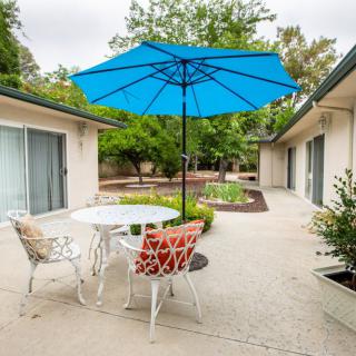 A cozy patio area with a blue umbrella, white metal table, and colorful chairs, surrounded by greenery.