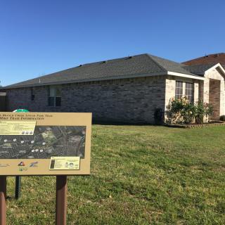 A suburban brick house with a grassy yard and a sign in the foreground under a clear blue sky.