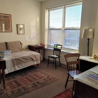 A cozy bedroom with a large window, various furniture items, and natural light streaming in.