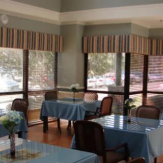 A brightly lit dining room with tables, chairs, and large windows allowing sunlight to filter in.