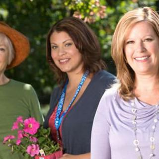 Three women smiling outdoors; one holding flowers in a garden setting.