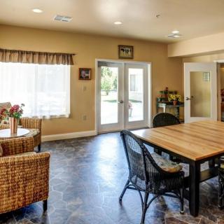 A cozy living room interior with wicker chairs, a wooden table, and ample natural light from large windows.