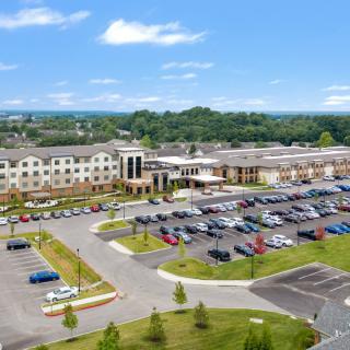 Aerial view of a large building complex with an adjacent parking lot filled with cars, surrounded by greenery and trees under a blue sky.