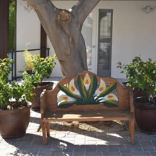 A decorative wooden bench with colorful detailing, surrounded by potted plants under a large tree on a patio.