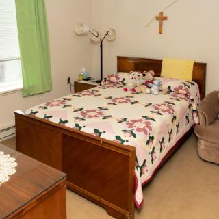 A cozy bedroom with a floral quilt, lamp, and a cross on the wall.
