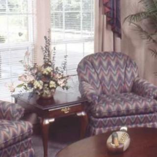 A cozy living room featuring patterned armchairs, a wooden table with a floral arrangement, and large windows with curtains.
