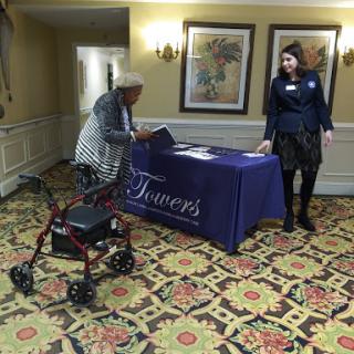 An elderly person with a walker approaches a registration table assisted by a staff member.