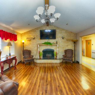 A cozy living room with hardwood floors, a fireplace, and a television mounted above. The space is furnished with armchairs and illuminated by a decorative chandelier.