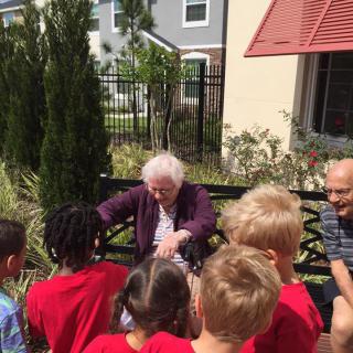 An elderly woman interacts with a group of children outside on a sunny day.