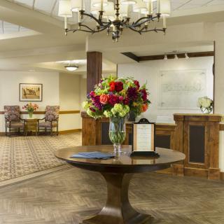 A hotel lobby with a centerpiece table adorned with vibrant flowers under an elegant chandelier.