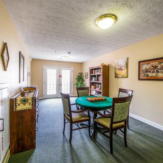 A cozy dining area with a table, chairs, and a bookshelf, illuminated by ceiling lights.