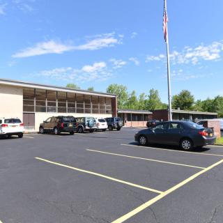 A parking lot in front of a large building with several parked cars and a flagpole on a clear day.
