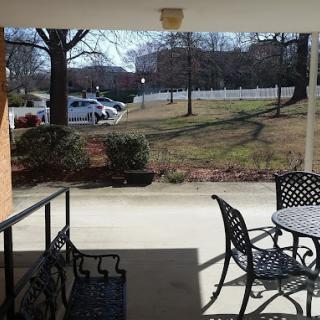 A view from a patio with metal chairs and a table, overlooking a grassy area with trees and a parking lot in the background.