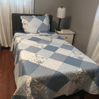A neatly made single bed with a quilted cover, next to a nightstand and lamp in a cozy bedroom.