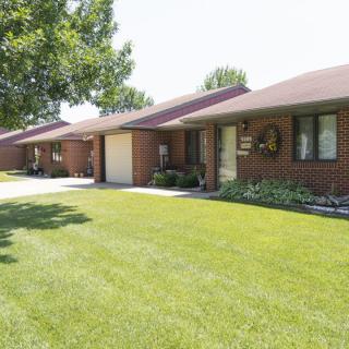 A suburban row of brick houses with well-maintained lawns and a tree along the sidewalk on a sunny day.