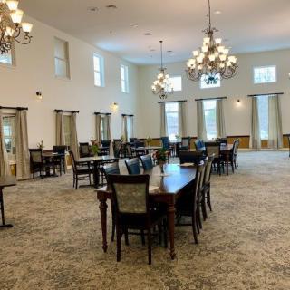 A spacious dining room with multiple tables and chairs, illuminated by elegant chandeliers and natural light from large windows.