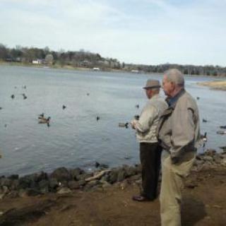 Two men fishing by a lake with ducks swimming nearby.