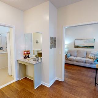 A cozy interior view featuring a living room and bathroom, connected by a hallway with a small vanity area. The space is adorned with simple home decor and hardwood floors.
