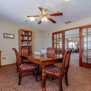 A cozy dining room with a wooden table and chairs, featuring a bookshelf and a ceiling fan, bordered by glass doors.