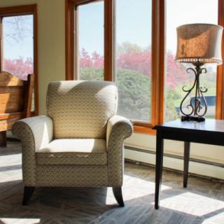A cozy armchair next to a small table with a lamp, bathed in natural light from large windows.
