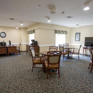 A spacious room with tables, chairs, a television, and office equipment, featuring carpeted flooring and natural light from windows.
