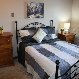 A cozy bedroom with a neatly made bed, flanked by wooden nightstands, each topped with a lamp and other decor. A winter-themed painting hangs on the wall above the bed.