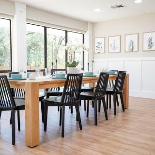A modern dining room with a large wooden table and black chairs, complemented by ample natural light from large windows.