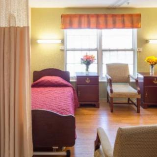 A brightly lit hospital room with a single bed, chairs, and floral decorations.