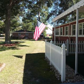 A sunny yard with a house displaying an American flag near a white picket fence.