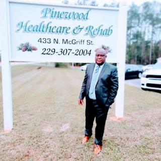 A man in a suit standing beside the Pinewood Healthcare & Rehab sign.