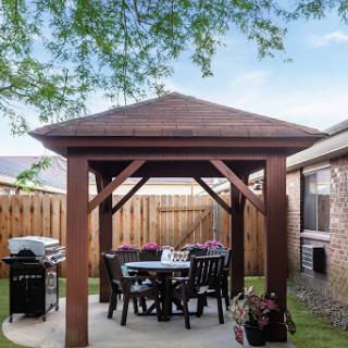 A cozy outdoor gazebo in a backyard, complete with dining furniture and a grill.