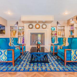 A vibrant living room featuring blue armchairs and a patterned rug, centered around a modern fireplace with surrounding bookshelves.