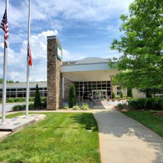 A modern building entrance framed by green grass and trees, showcasing flagpoles under a clear blue sky.